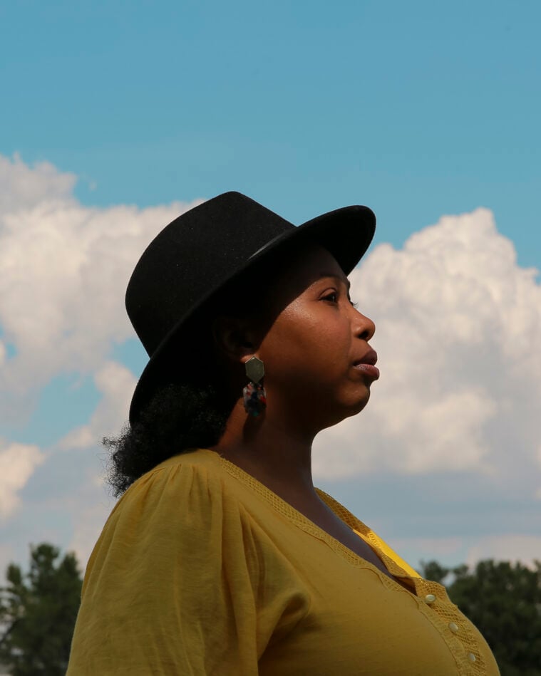 Deborah Mouton is a Black woman in a black Stetson hat, wearing a yellow-green scoop neck dress. She is outdoors, profiled against a treeline and a partly cloudy sky.